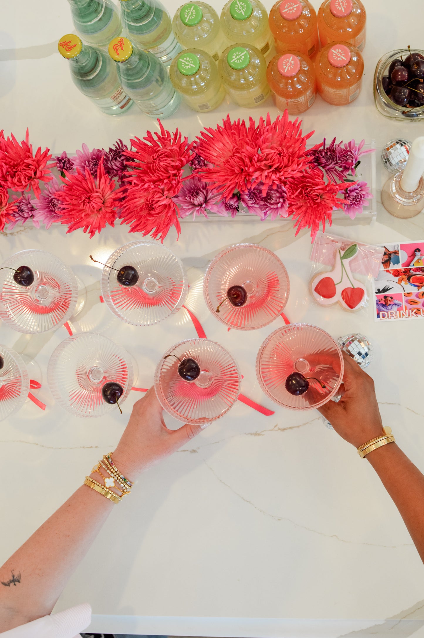 Clean energy cocktail setup with pink drinks, flowers, and bottles on a white marble surface.